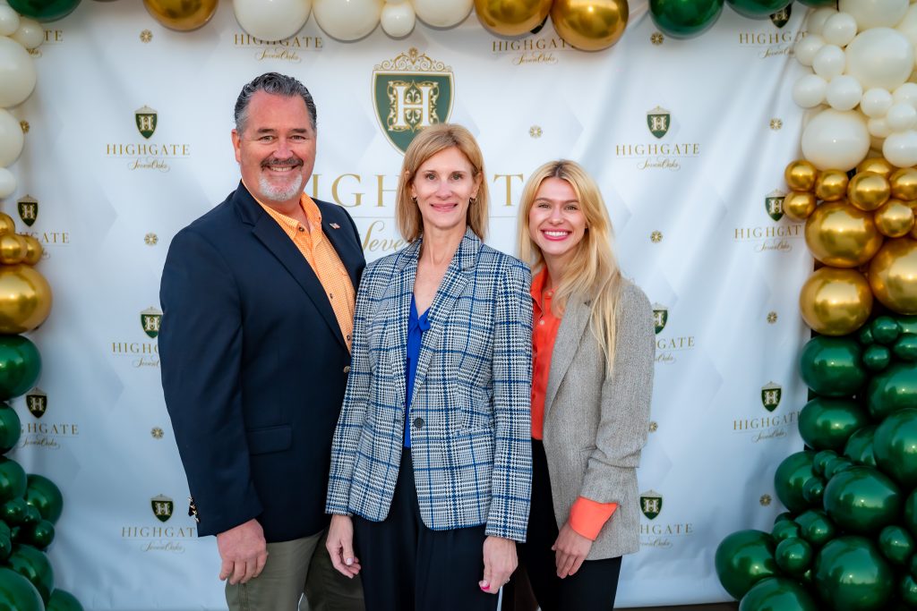 Three people stand and smile in front of a backdrop and balloon arch with green, gold, and white balloons. The backdrop reads "Highgate" with a crest and gold lettering. All wear business attire.