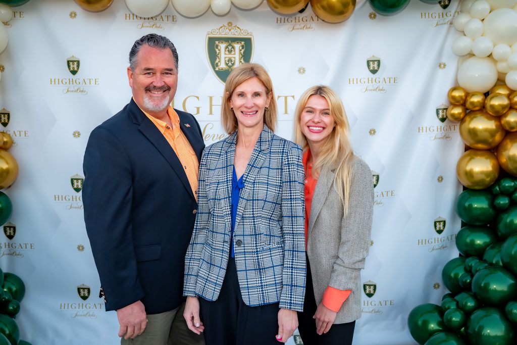 Three people stand smiling in front of a decorative backdrop with gold, white, and green balloons and a "Highgate" logo. Two women and one man are dressed in business attire.