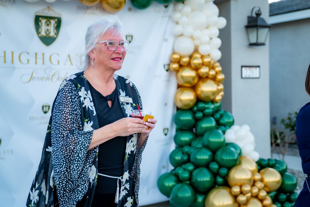 An older woman with white hair and glasses holds a drink while standing in front of a green, gold, and white balloon arch and a Highgate event backdrop outdoors.