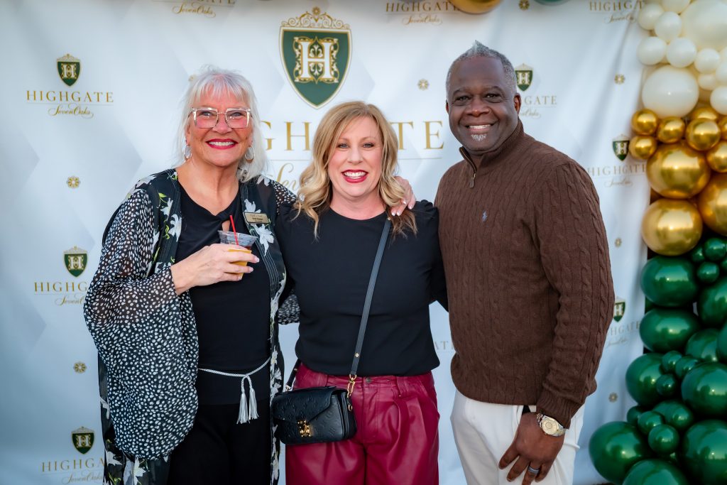 Three people stand smiling together at an event in front of a Highgate-branded backdrop and a gold, white, and green balloon arrangement. The group includes two women and one man, posing closely for the photo.