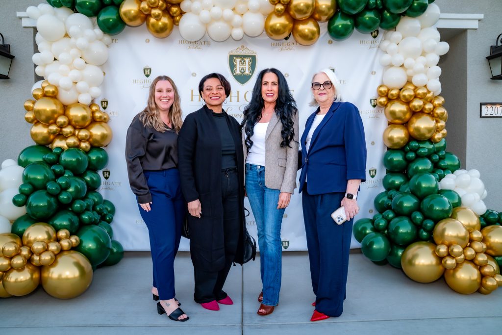 Four women pose and smile in front of a decorative backdrop with gold, white, and green balloons at an event. The backdrop features the Highgate logo and shield emblem.