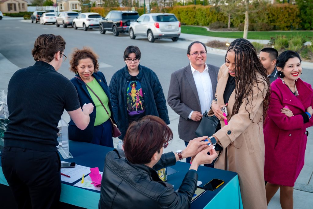 A group of people, some smiling and chatting, gather around an outdoor check-in table on a suburban street. One person hands a smartwatch to a woman, while others wait in line. Cars and houses are visible in the background.