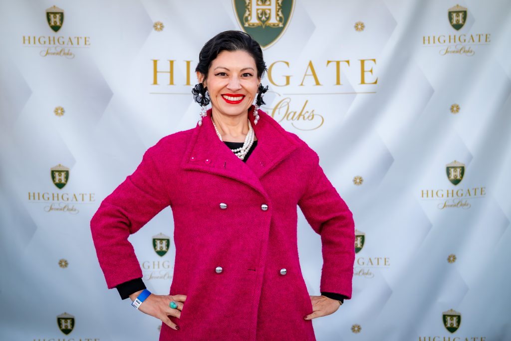 A woman in a bright pink coat smiles confidently with hands on hips, standing in front of a step-and-repeat banner that reads “Highgate” with gold logos.