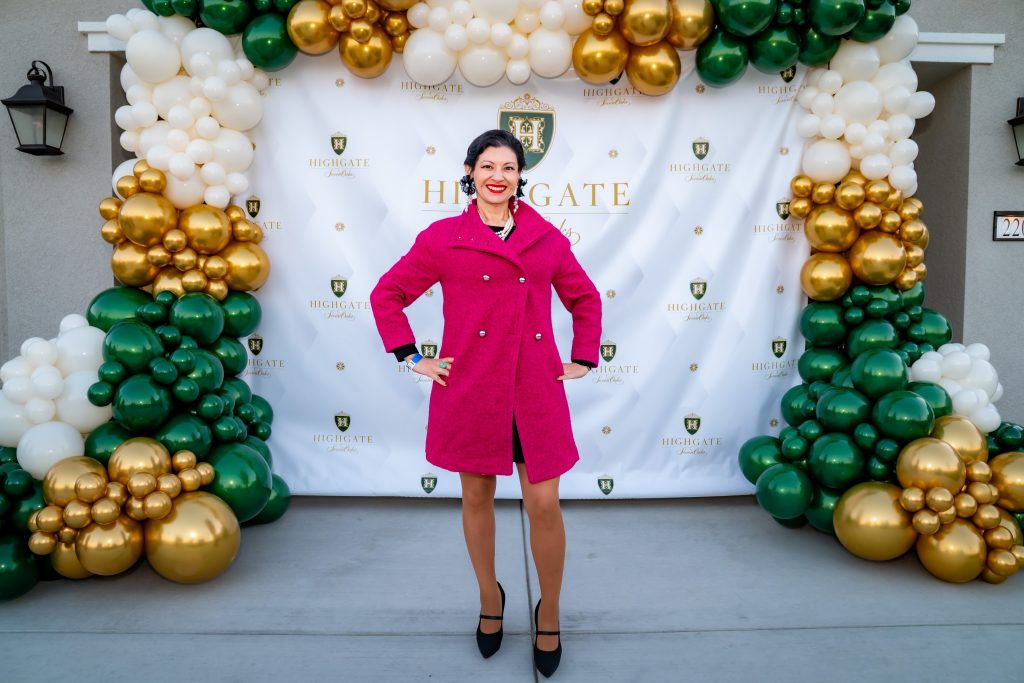 A smiling woman in a bright pink coat poses in front of a decorated backdrop with gold, green, and white balloons and the "Highgate" logo. She stands with hands on her hips on a gray pavement.