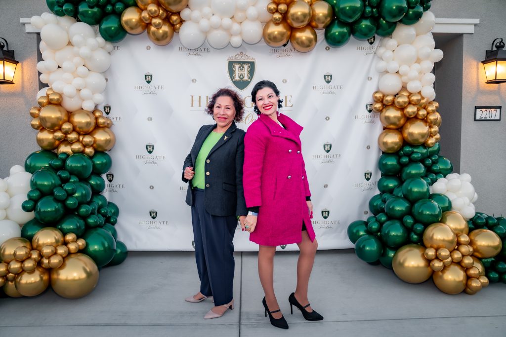 Two women pose and smile in front of a festive backdrop decorated with green, gold, and white balloons. One wears a dark blazer and pants, the other a bright pink coat. The backdrop reads "Highgate" with a crest logo.