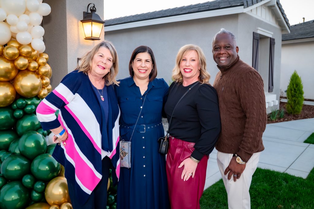 Four adults—three women and one man—stand smiling together outdoors near a house, with a decorative cluster of white and gold balloons to the left. It appears to be late afternoon or early evening.