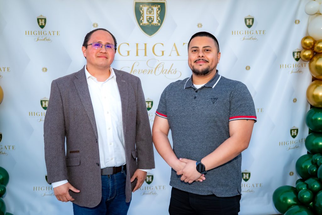 Two men stand in front of a white backdrop with gold and green "Highgate Seven Oaks" logos, smiling at the camera. One wears a blazer and jeans; the other wears a gray polo shirt. Balloon decorations are seen at the sides.