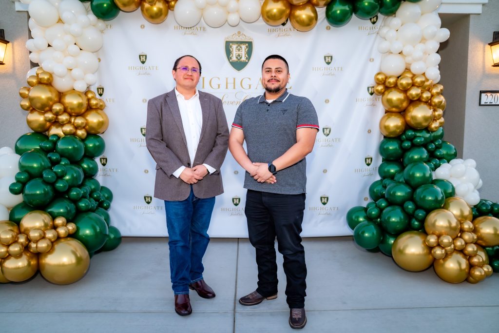 Two men stand in front of a festive balloon arch with white, green, and gold balloons and a “Highgate” backdrop, posing for a photo at an outdoor event.