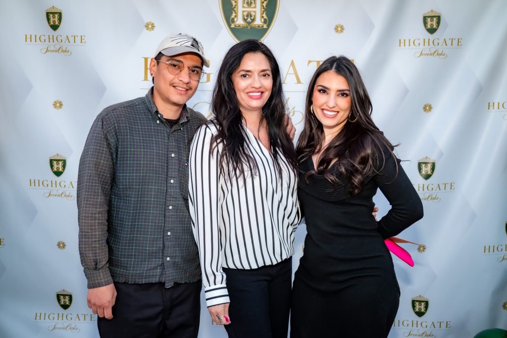 Three people stand smiling in front of a branded step-and-repeat backdrop with "Highgate" logos. The person on the left wears a cap and glasses, the middle person wears a striped blouse, and the person on the right wears a black dress.
