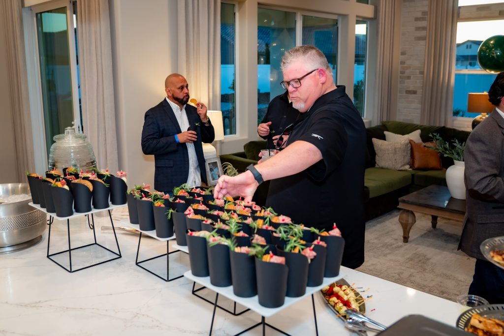 Two men are serving themselves food from a buffet table with cups filled with salad or appetizers in a modern, well-lit living room during a social event.
