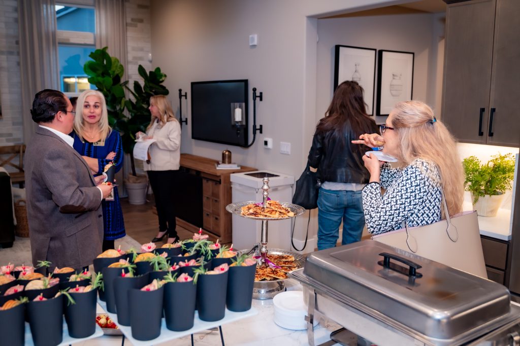 A group of people socialize in a modern kitchen during a gathering. A buffet with snacks and desserts is set up in the foreground, while guests chat and eat in the background.