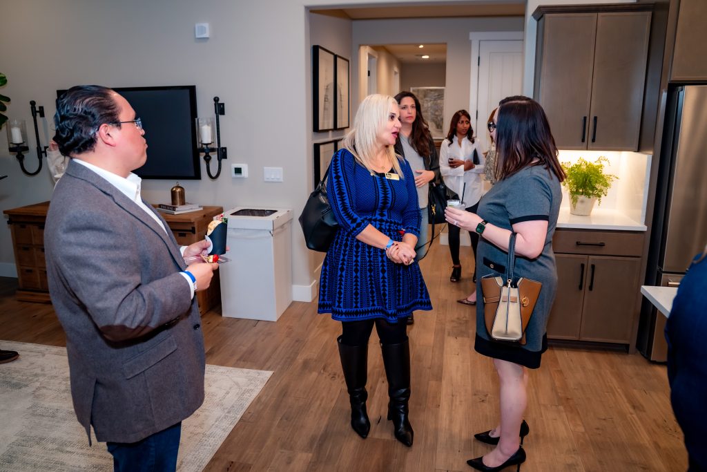 A group of people are talking in a modern kitchen and living space; two women stand conversing in the center, while others interact in the background. Everyone is dressed in business casual attire.
