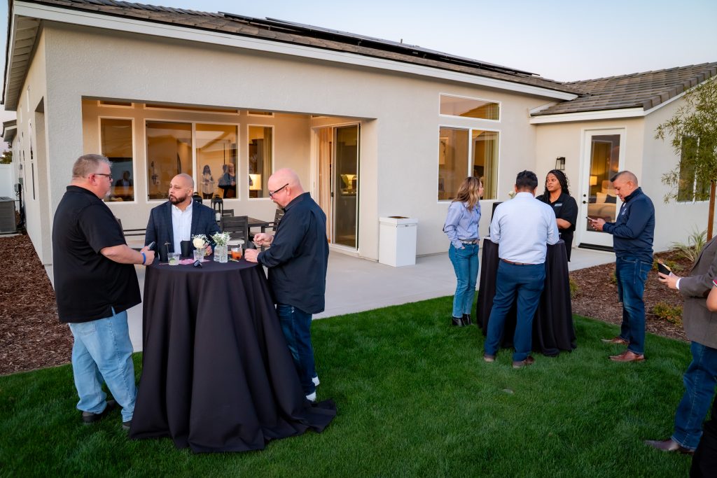 A group of people socializes in small groups around tables covered with black cloths in the backyard of a modern house during an outdoor gathering.