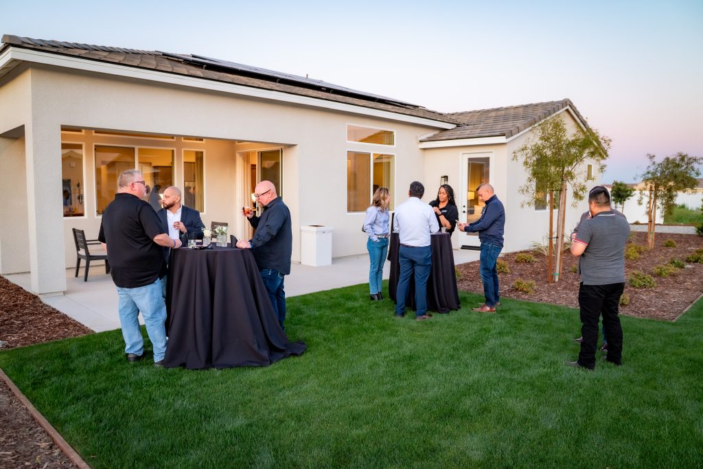 A group of people gather in small groups, talking and drinking around cocktail tables on a green lawn outside a modern house at sunset. The house has large windows and landscaped garden beds.