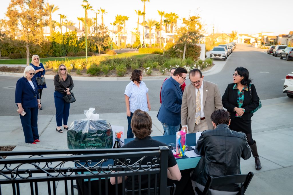 A group of people gathers outdoors near a table at the entrance of a residential area; some are checking in at the table while others wait or talk nearby, with palm trees and parked cars in the background.