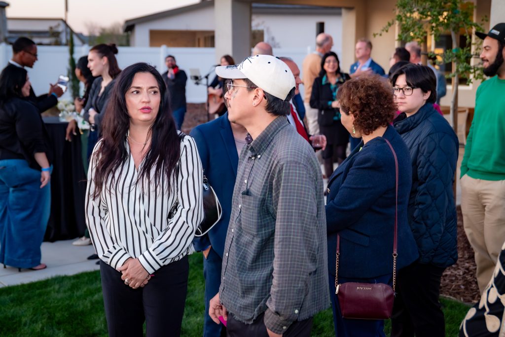 A group of people stand and talk at an outdoor gathering. Some are engaged in conversation, while others look around. The background features a house, trees, and more attendees.
