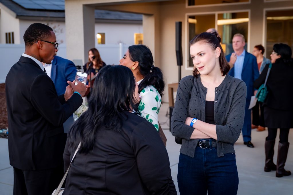 A group of people converse at an outdoor social gathering. A woman with crossed arms stands in the foreground, while others chat in pairs in the background near a building. The setting appears casual and friendly.