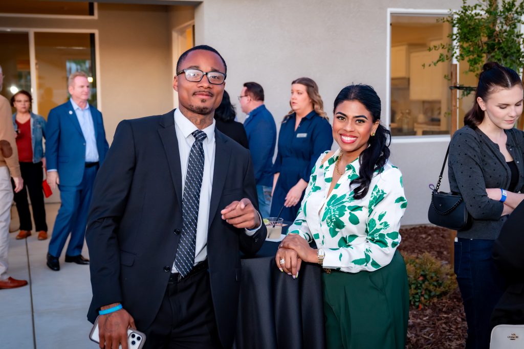 Two people stand smiling at an outdoor event, dressed in business attire. The man wears a suit and tie, and the woman wears a white blouse with green floral prints. Other attendees mingle in the background.