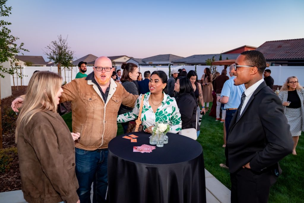 A group of people socialize outdoors at a formal event, standing around a table with a black tablecloth and flowers. More guests are mingling in the background as the sun sets.