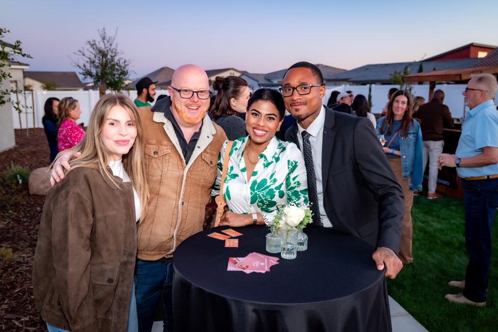 Four people stand smiling around a tall black table with flowers and cards at an outdoor event; other guests and houses are visible in the background during sunset.