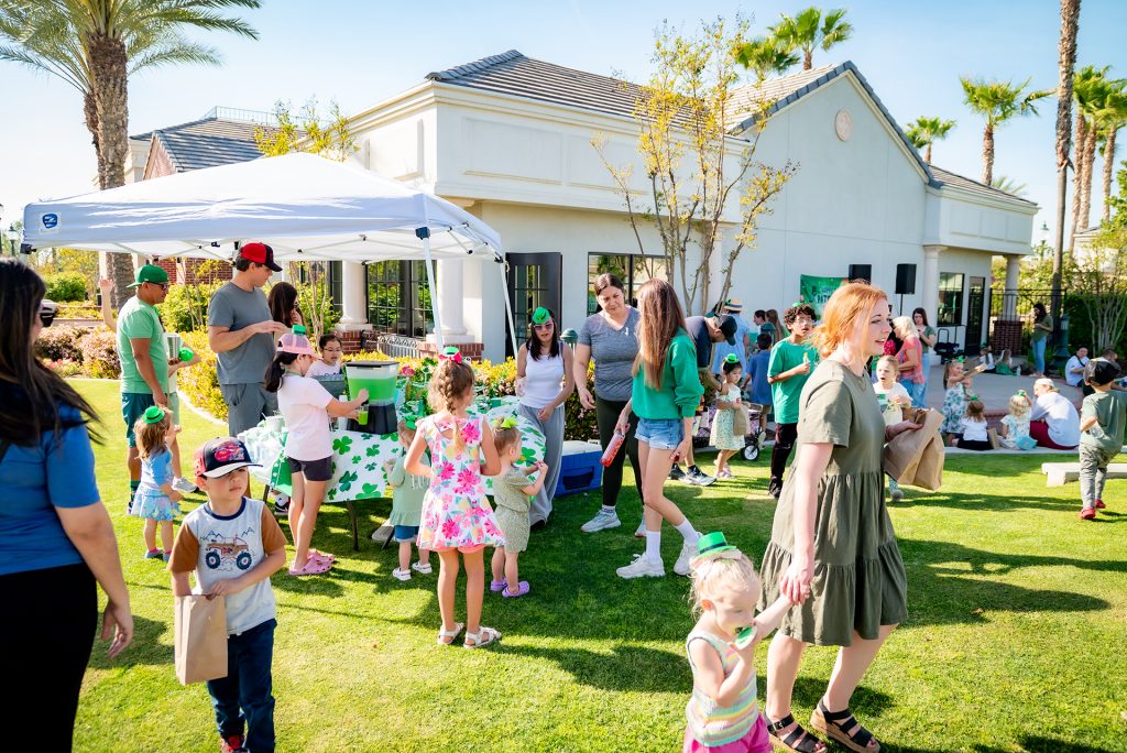 A group of adults and children enjoy a sunny outdoor gathering with games, food, and decorations, including a white canopy, in a grassy yard in front of a white house. Many people are wearing green outfits.