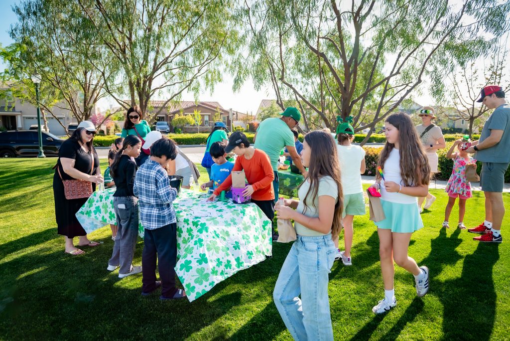 Children and adults gather around a table covered with a shamrock-patterned cloth at an outdoor event in a sunny park. Some kids hold baskets while others receive items from people behind the table. Trees and houses are visible in the background.