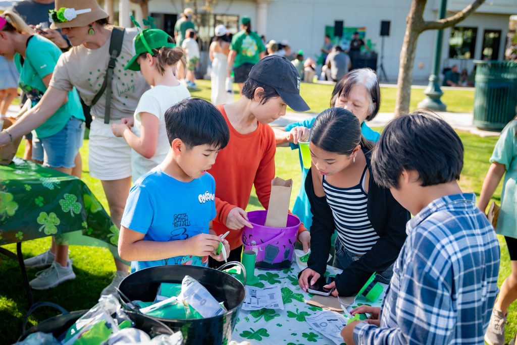 Children and adults gather around a table outdoors, engaging in crafts and activities with green shamrock decorations. People are focused and interacting, enjoying a sunny community event.