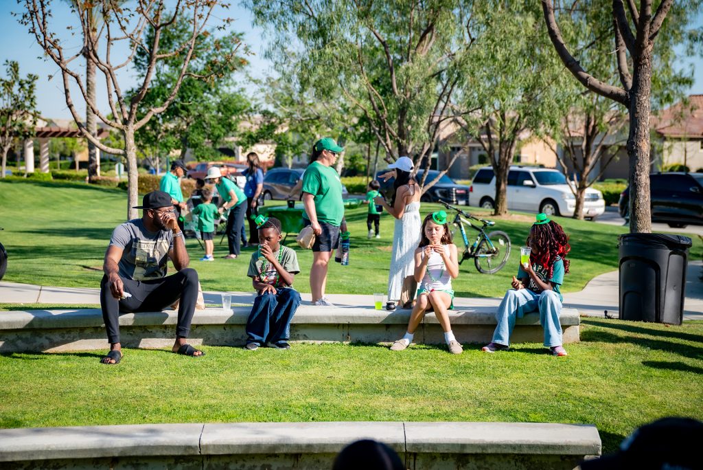 A group of people sit and stand on grass in a sunny park. Four people sit on a low wall in the foreground, while others gather and talk in the background. Trees and parked cars are visible.