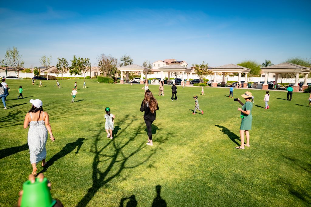A group of children and adults are playing and walking on a large grassy field in a park on a sunny day, with houses and gazebos visible in the background.