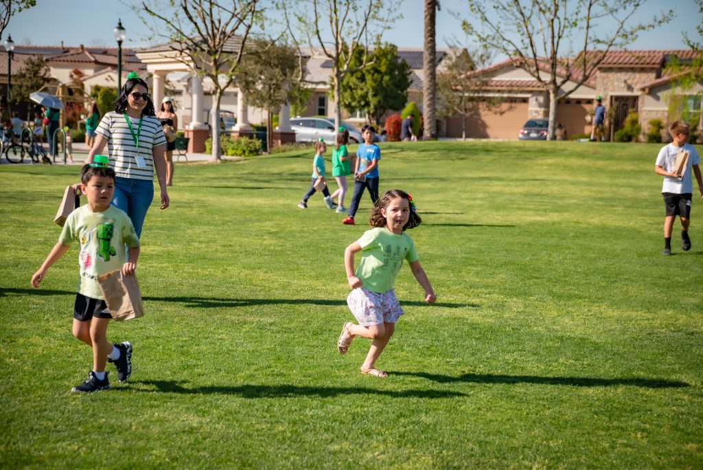 Children run and play on a grassy field on a sunny day, while a woman watches them in the background. Other kids and adults are scattered around the park, enjoying the outdoors. Trees and houses line the background.