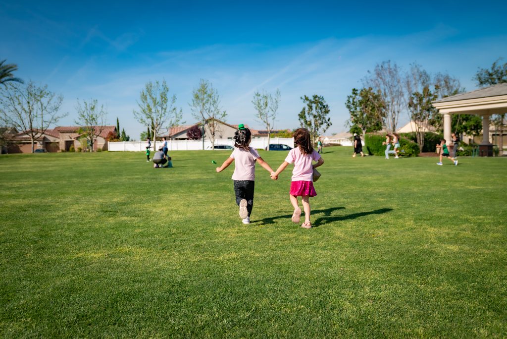 Two young girls holding hands run across a grassy park under a clear blue sky, with trees, houses, and other people in the background enjoying the sunny day.