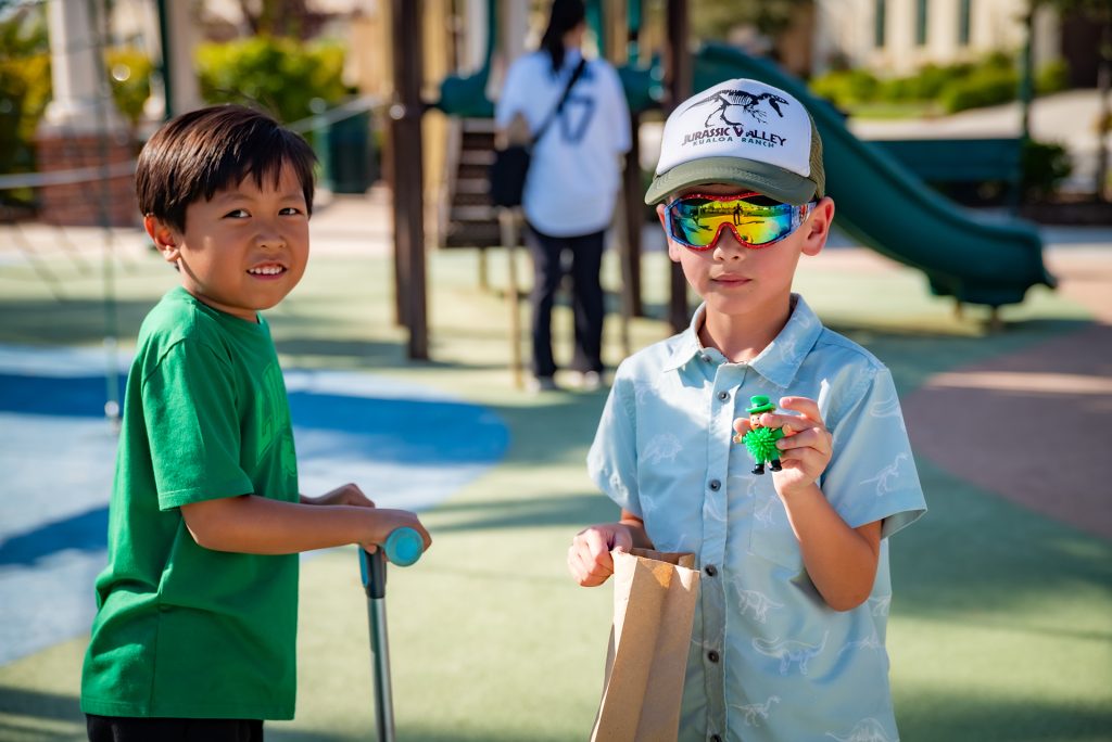 Two young boys stand at a playground; one holds a scooter and smiles, while the other wears a cap and sunglasses, holding a paper bag and a small toy dinosaur. Playground equipment and another child are in the background.