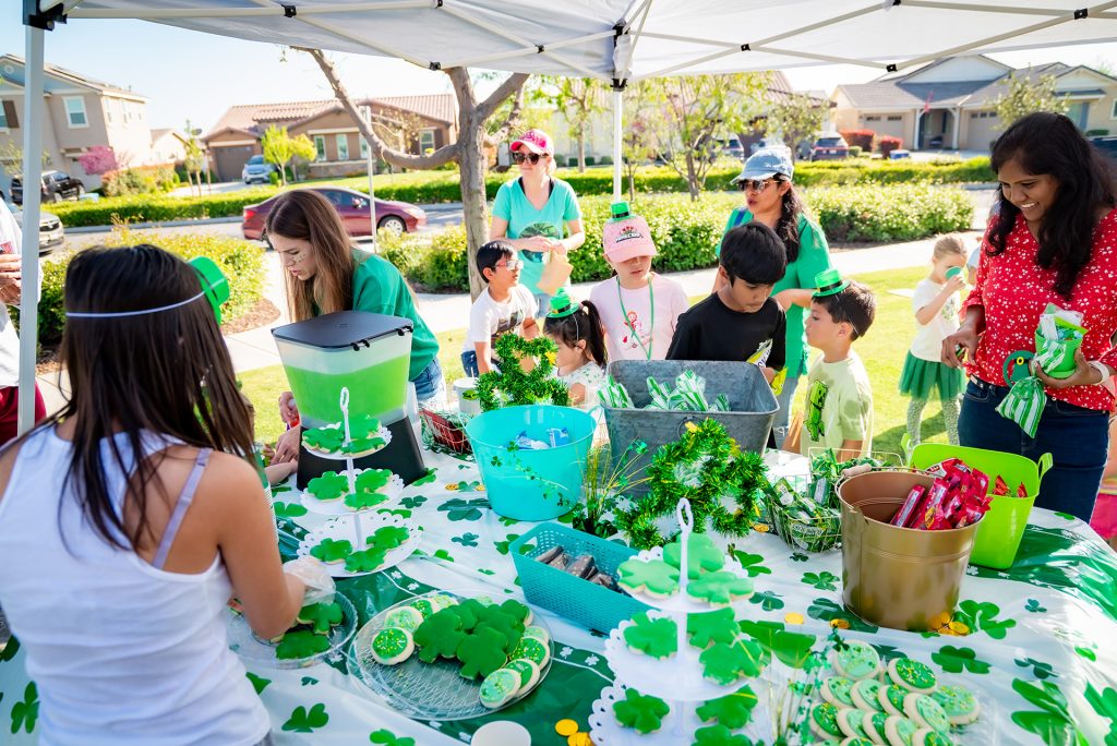 A group of adults and children, many dressed in green, gather around a festive outdoor table decorated with shamrocks, green treats, and drinks, celebrating St. Patrick’s Day in a suburban neighborhood.