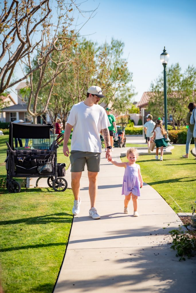 A man wearing a white shirt, shorts, and a cap walks hand-in-hand with a young girl in a purple dress on a sunny park path, with people and trees in the background and a stroller nearby.