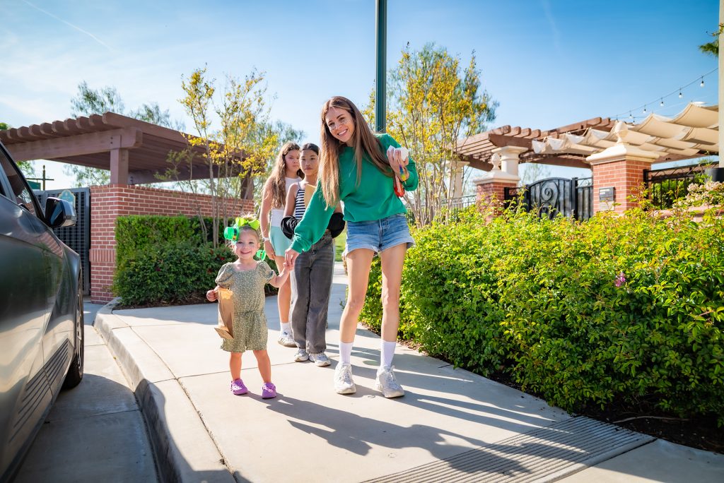 A smiling woman in a green shirt holds hands with a small child in a dress and green bow, walking on a sunny sidewalk with two other people and a car nearby. Trees and brick structures are in the background.