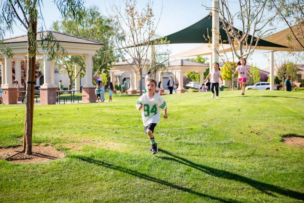 A young boy in a white jersey runs across a grassy park, with other children running behind him. Trees, pavilions, and people are visible in the background on a sunny day.