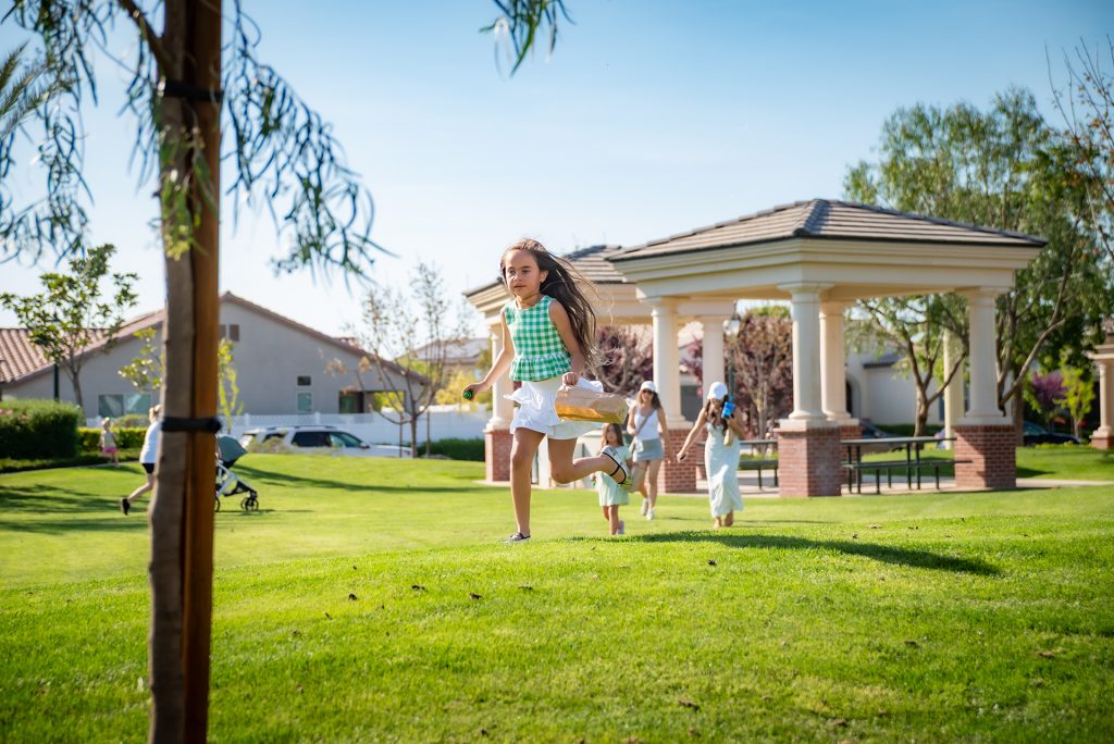 Three children run and play on green grass in a sunny park with a pavilion in the background. Trees, houses, and a stroller are visible, creating a lively, outdoor scene.