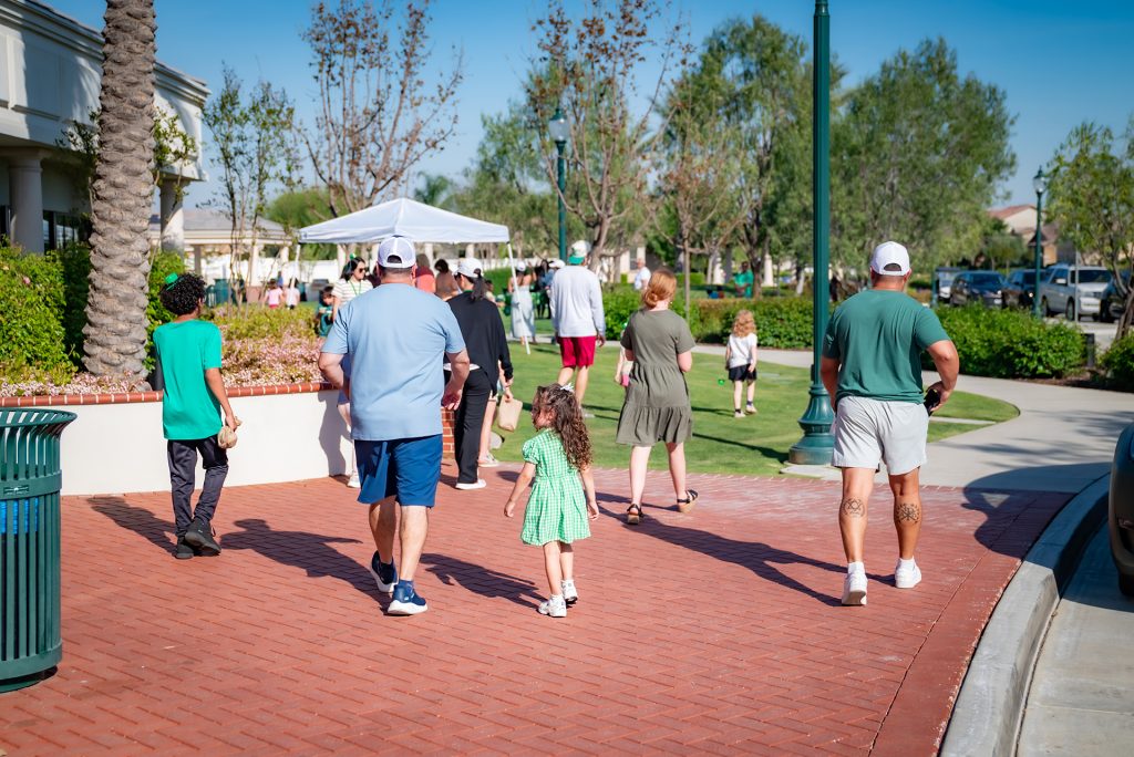 People of various ages walk along a red brick path toward a park with green grass and trees. Some adults wear casual clothes and hats, and a young girl in a green dress walks beside them. A canopy and parked cars are visible ahead.
