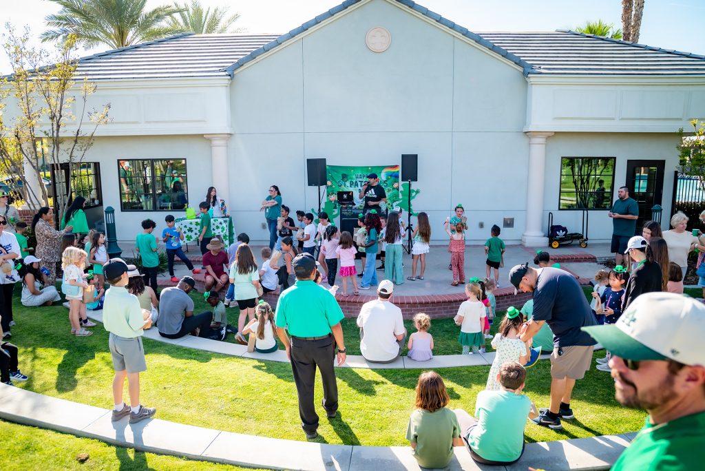 A crowd of people, including many children, gather outdoors in front of a building for a festive event with green decorations. Some children are on stage, and others sit or stand on the grass, watching the performances.