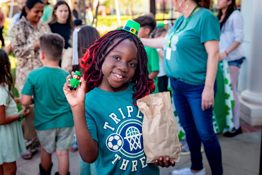 A smiling girl with red braids, wearing a green "Triple Threat" shirt and a small green hat, holds a brown paper bag and a green toy. People are gathered around her, some wearing green for a festive outdoor event.