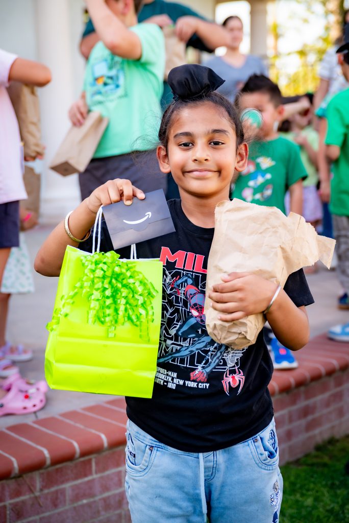 A smiling child stands outside holding a neon green gift bag, a small black envelope, and a brown paper bag. Other children are visible in the background, some holding bags, at what appears to be a party or gathering.