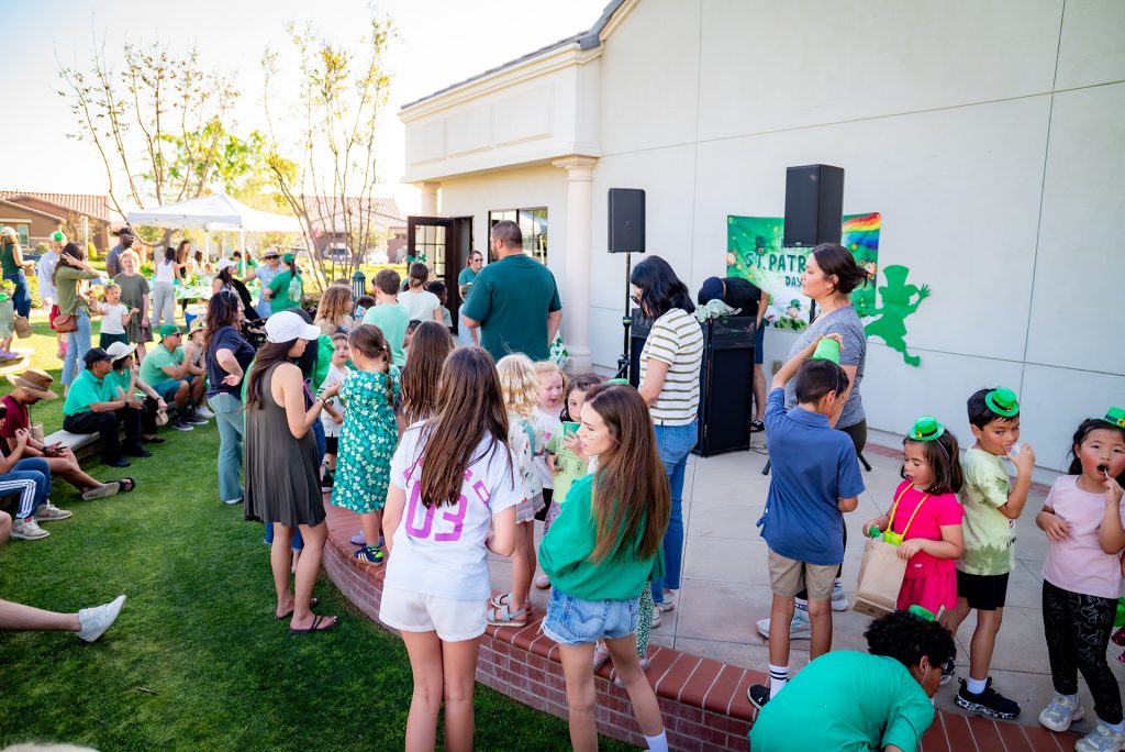 A group of children and adults, many wearing green, gather outdoors near a building decorated for a St. Patrick's Day event. Some kids have face paint and festive headbands, while others chat or play on the grass.