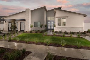 A modern, single-story house with clean lines, gray and white exterior, large windows, double front doors, and a well-maintained yard featuring green grass, small shrubs, and a concrete walkway at sunset.