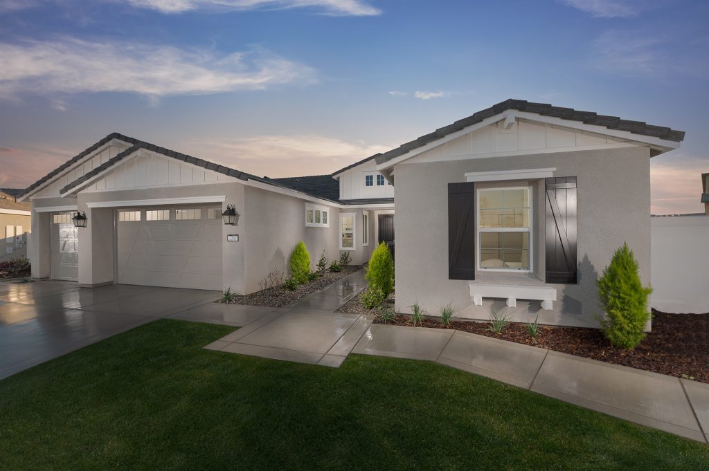 Modern single-story house with light gray exterior, dark roof, and white trim. Large windows, garage with double doors, small bushes and neatly landscaped yard. Evening sky with soft clouds in the background.