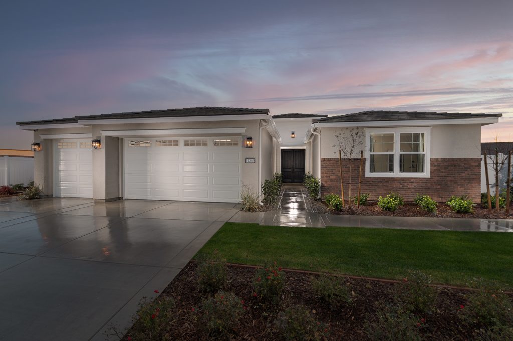 Single-story modern house with three white garage doors, large windows, a double front door, and a neatly landscaped front yard, taken at sunset with a wet driveway reflecting the soft evening light.