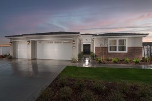 Single-story modern house with three white garage doors, large windows, a double front door, and a neatly landscaped front yard, taken at sunset with a wet driveway reflecting the soft evening light.