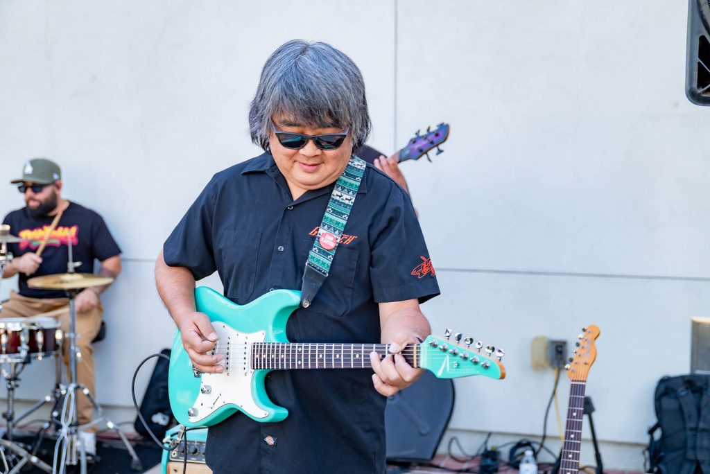 A man with gray hair and sunglasses plays a turquoise electric guitar outdoors. A drummer and bassist perform in the background against a white wall. The mood appears casual and relaxed.