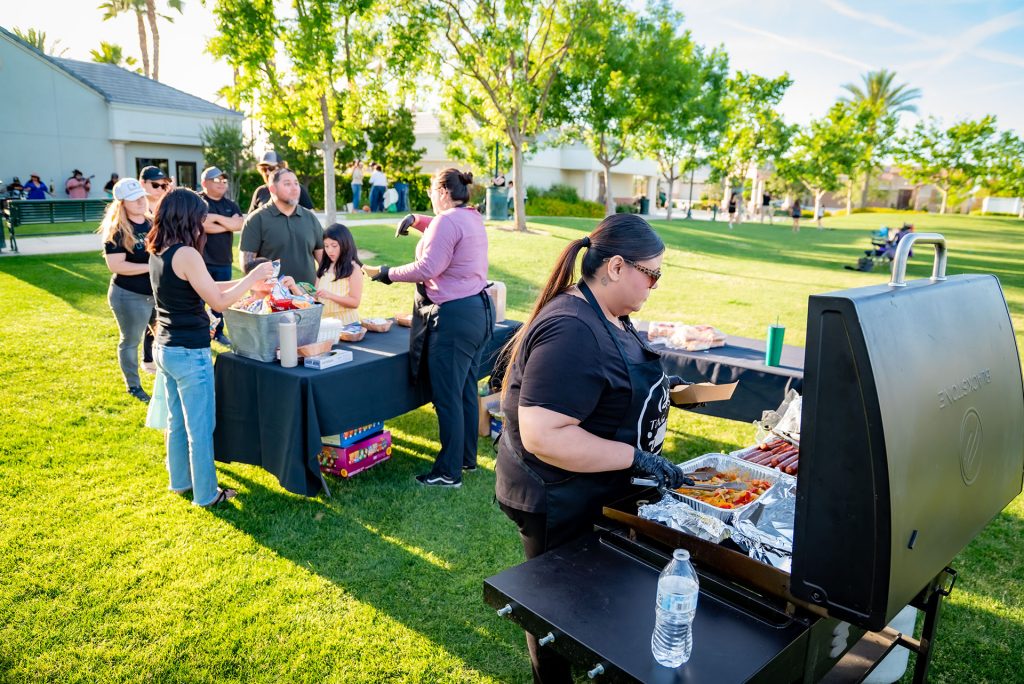People gather outdoors on a sunny day, some serving food from a grill and table with snacks and drinks. Trees, grass, and buildings are visible in the background, creating a relaxed picnic atmosphere.