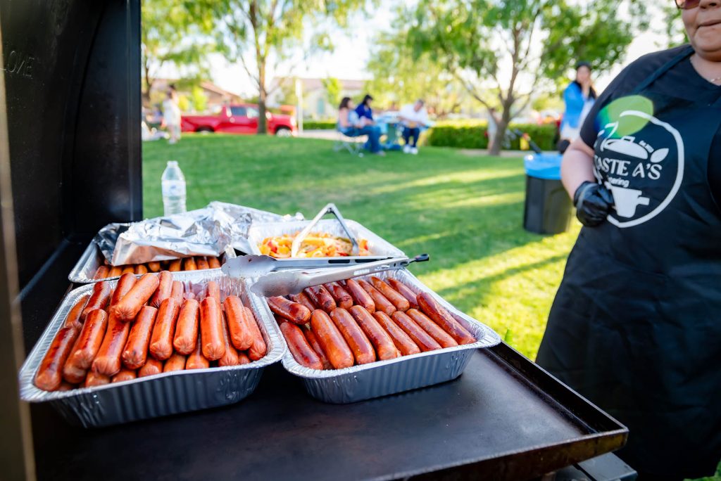 Three trays of grilled hot dogs sit on a barbecue grill. A person in a black apron stands nearby. In the background, people are seated on a green lawn with trees and sunlight.