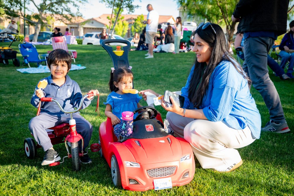 A woman smiles while kneeling beside two young children sitting in toy cars on grass, sharing snacks at a park with other families in the background on a sunny day.
